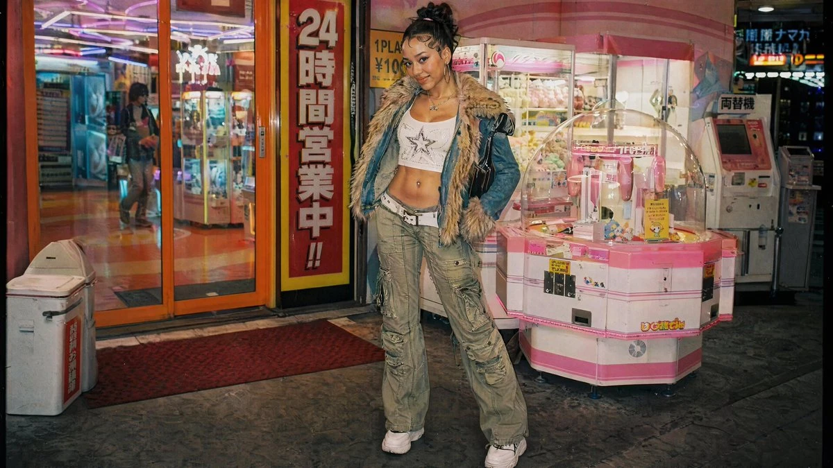 A young woman poses confidently in a trendy outfit, featuring a cropped top and cargo pants, against a vibrant arcade backdrop
