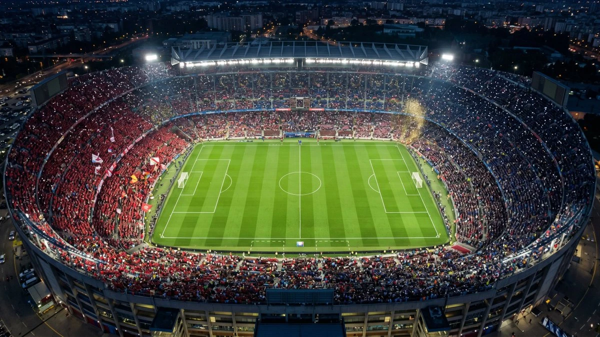 Aerial night view of a packed soccer stadium with bright floodlights, green pitch and red-blue crowds