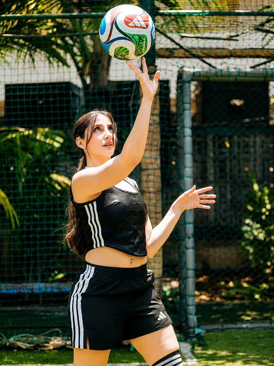 A young athletic woman in a black sports outfit tosses a colorful soccer ball upward on a sunny outdoor court.