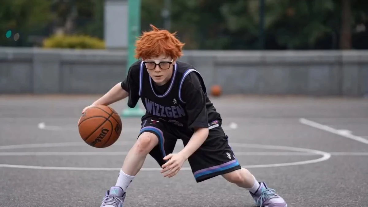 A young person with short orange hair and glasses dribbles a basketball on an outdoor court, wearing a black jersey and shorts