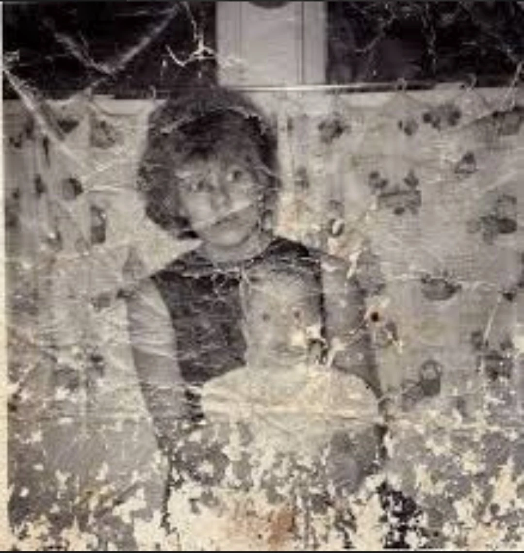A heavily damaged black-and-white portrait shows a woman in a dark dress holding a toddler against her chest, with a patterned curtain in the background