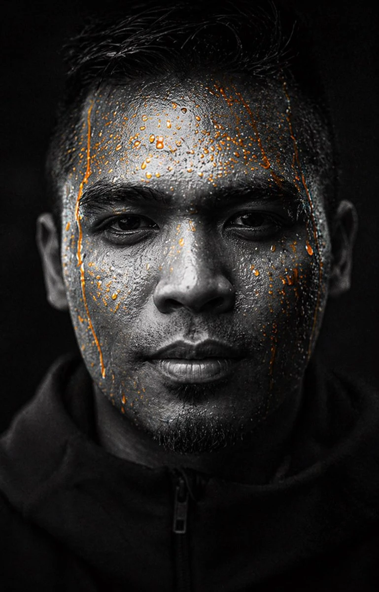 A male athlete with short dark hair and stubble stares intensely at the camera, illuminated by low-key lighting against a nearly black background, with droplets of orange liquid