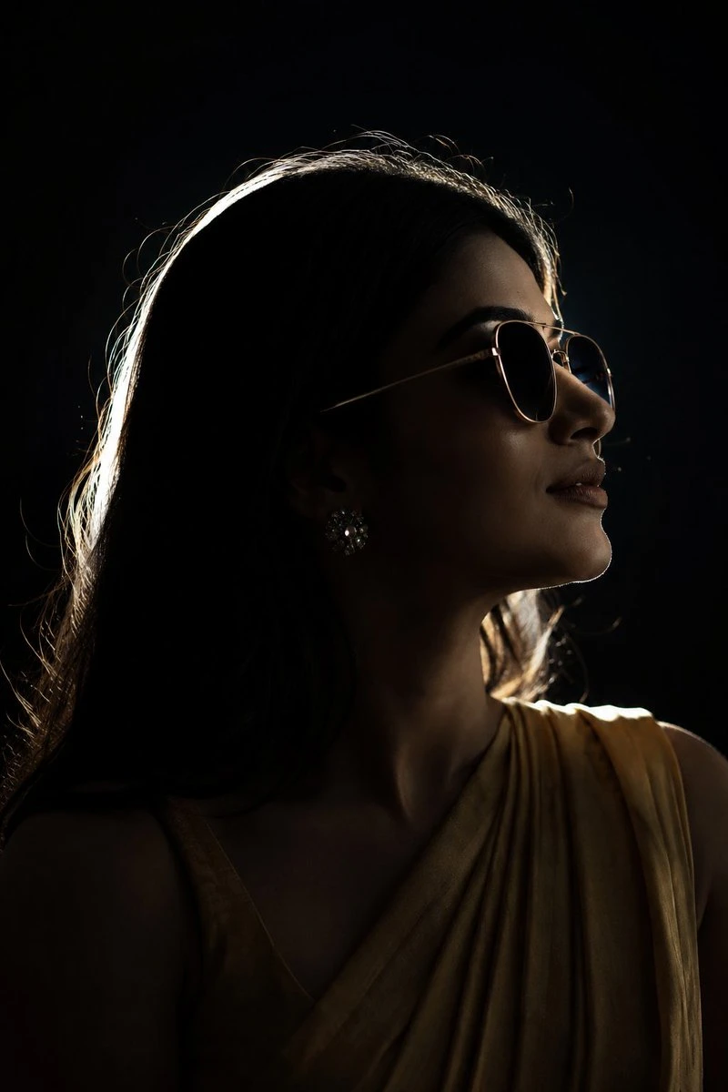 A stylish young woman in sunglasses is captured in a dramatic side profile, illuminated by rim lighting against a deep black background
