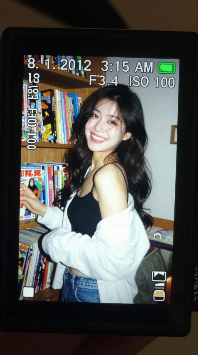 A close-up of a digital camera screen displays a candid photograph of a young woman laughing beside a shelf filled with colorful comic books