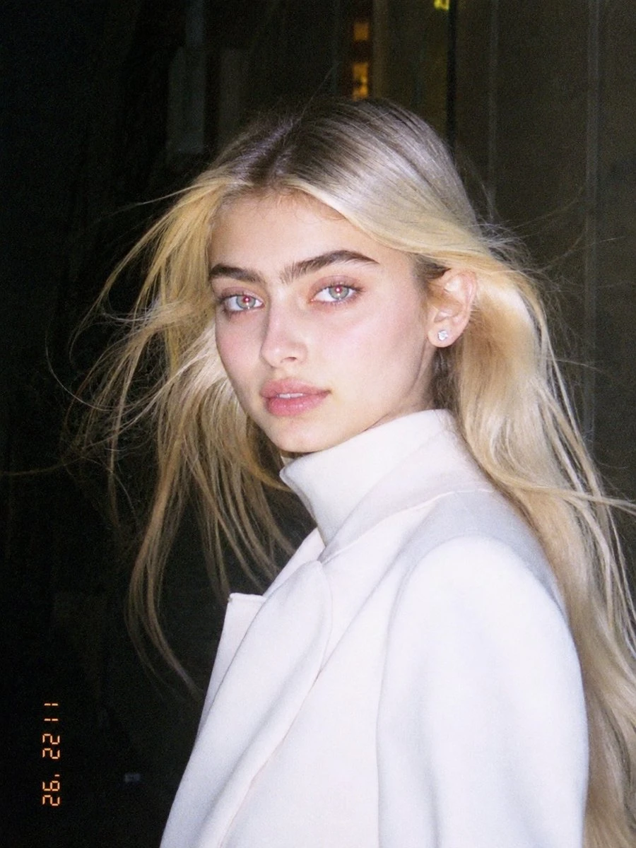 A young woman with long blonde hair poses in a white coat, captured with harsh flash highlights and a nostalgic, low-resolution aesthetic
