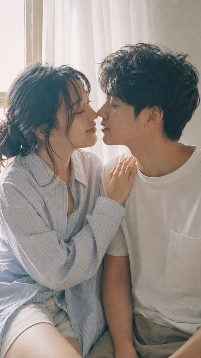 A young Japanese couple shares an intimate moment, leaning close together in soft pastel tones with natural daylight filtering through a window