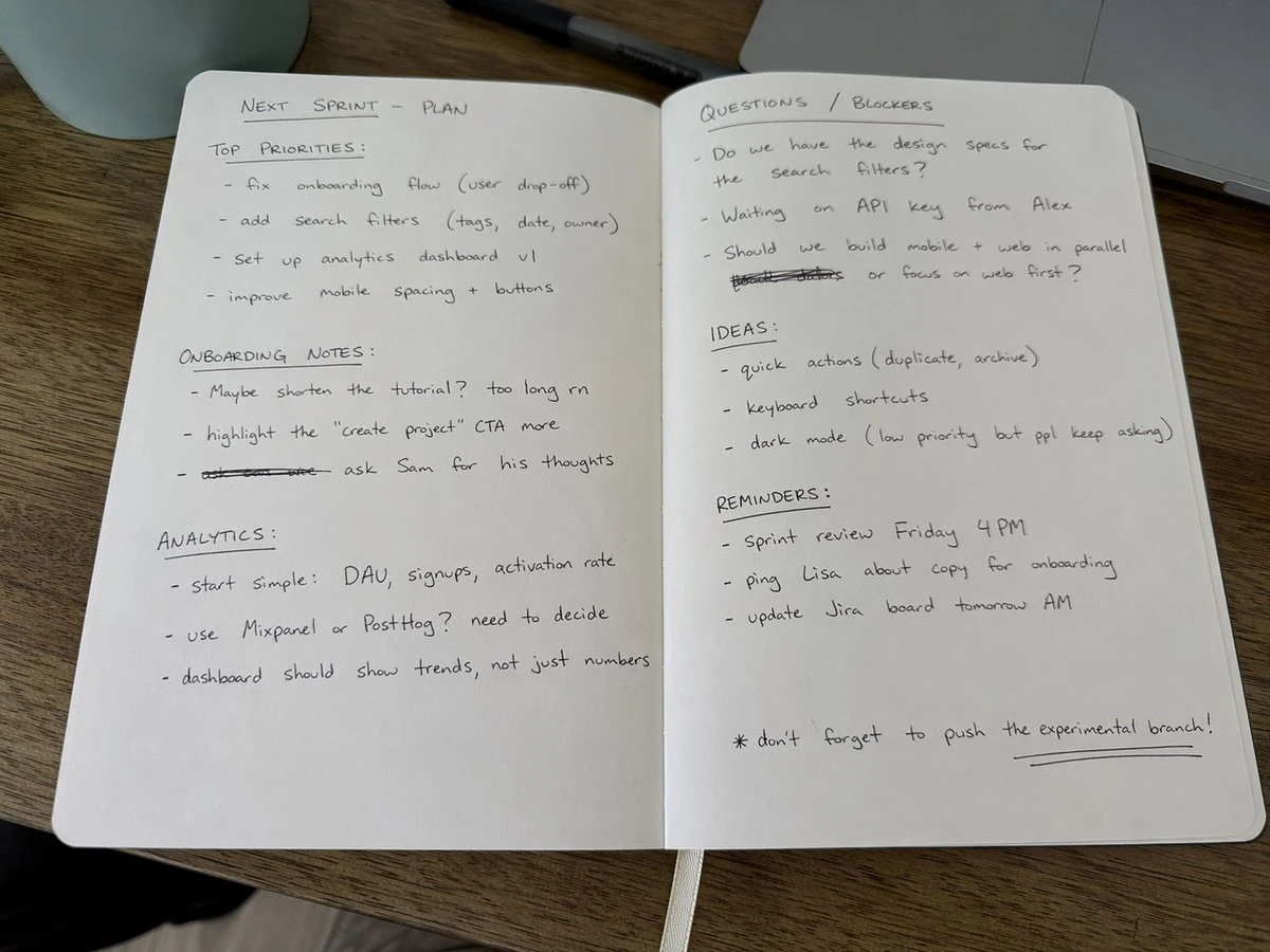 An open notebook with casual handwritten notes in black ink lies flat on a wooden desk, illuminated by natural daylight