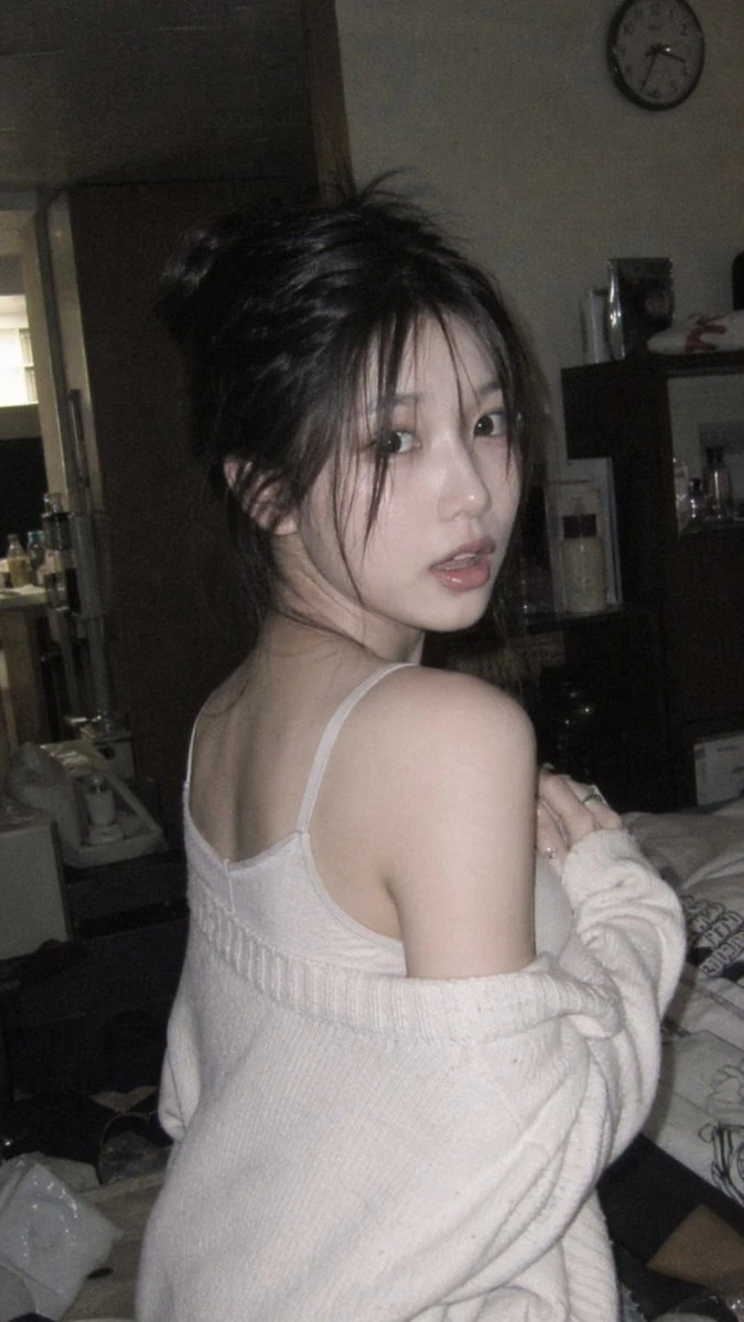 A person with long hair is seen from the side, wearing a light top and a sweater, in a softly lit room.