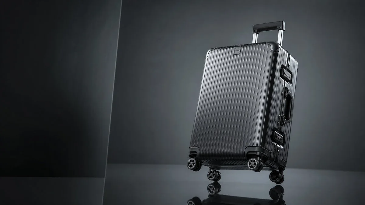 A premium hard-shell rolling suitcase in glossy gunmetal black features vertical ribbed grooves and four double-spinner wheels, positioned at a slight angle.