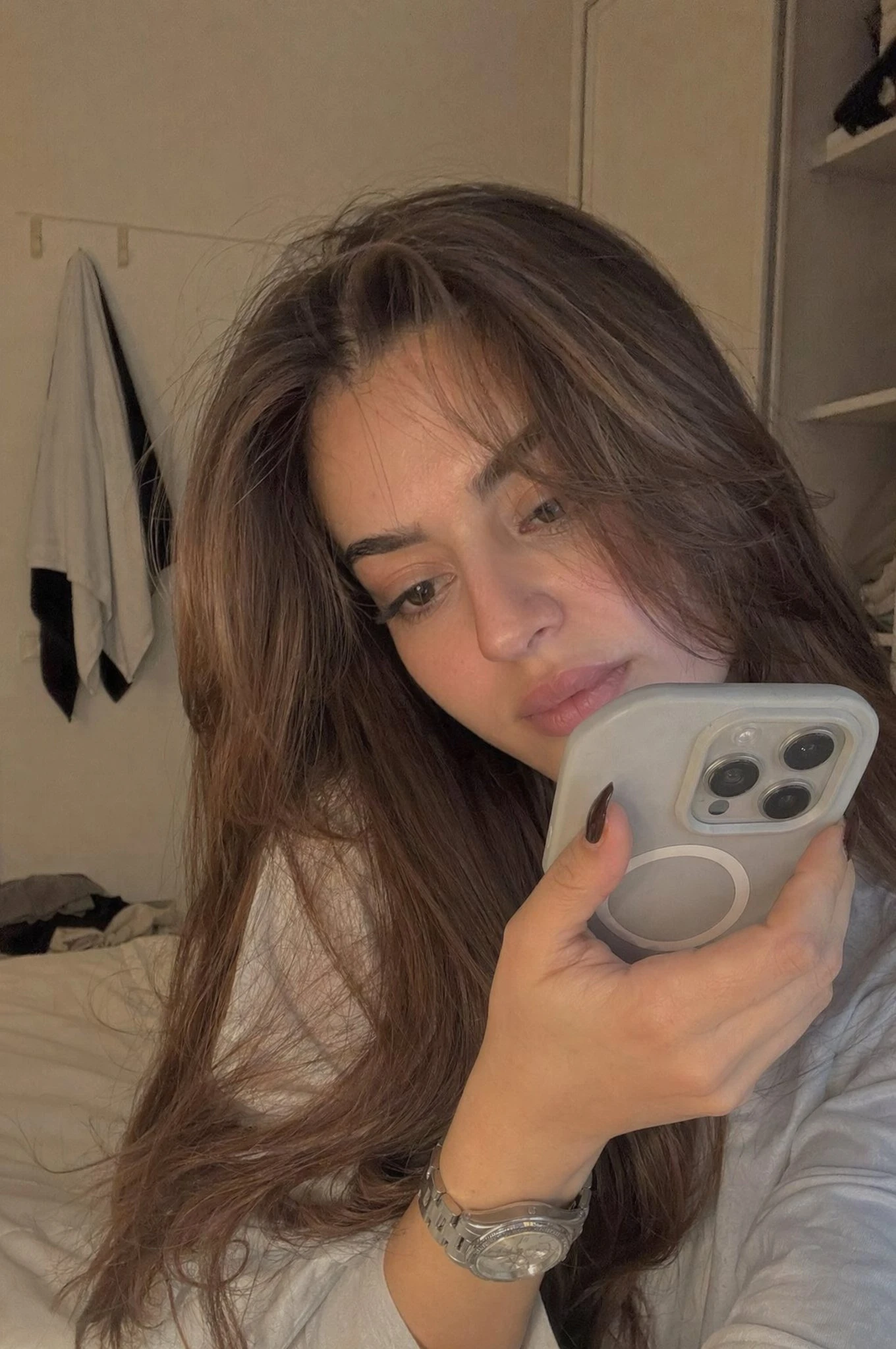 A young woman with long hair sits in a softly lit bedroom, holding her phone close as she records a private video, exuding a thoughtful expression