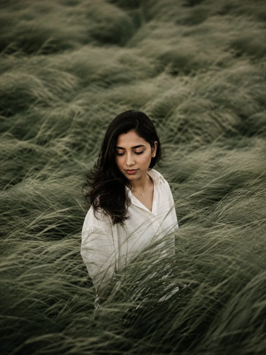 Soft editorial portrait shows a woman in a white blouse standing among tall sage-green grasses
