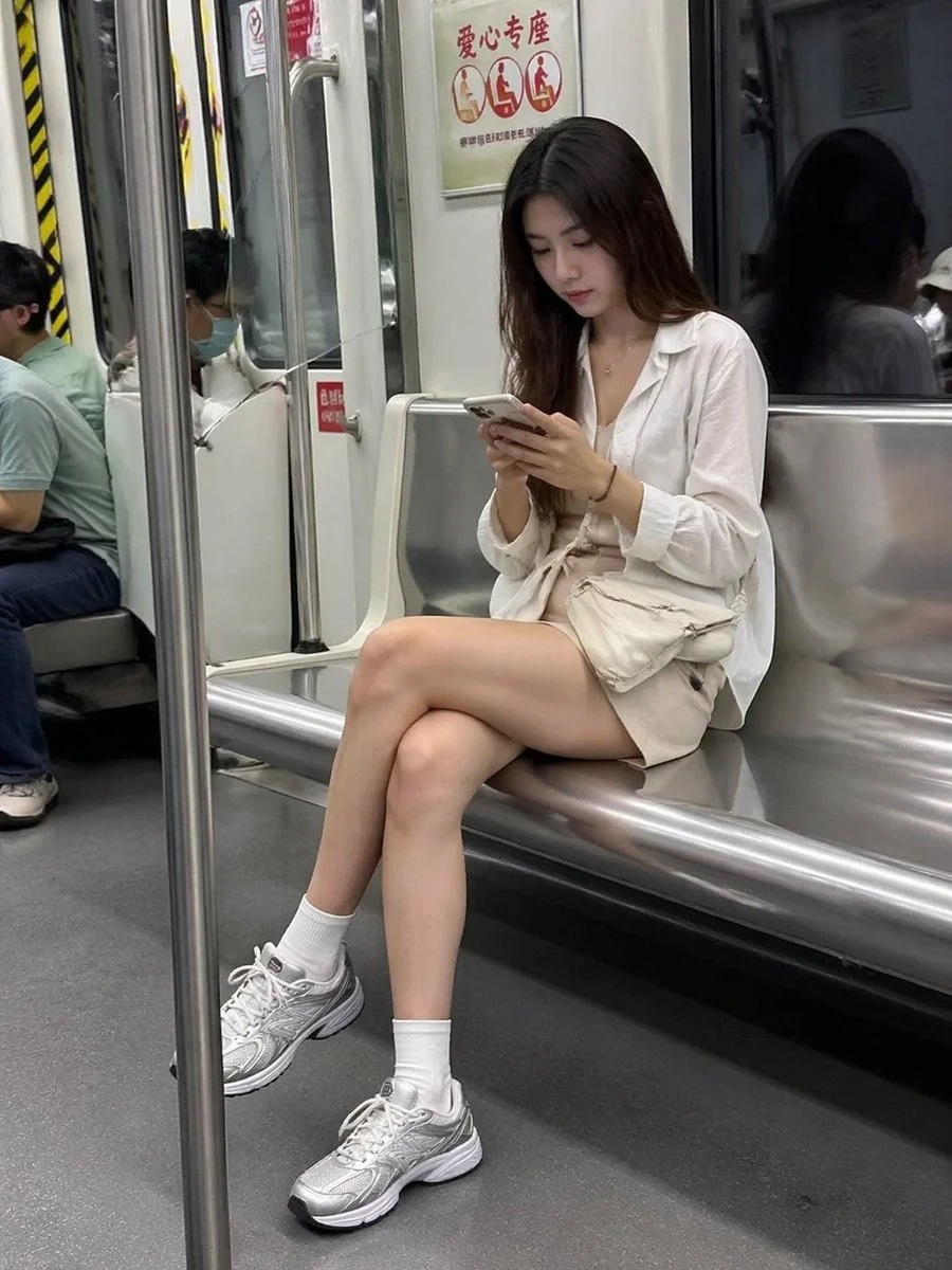 A woman in a white blouse and shorts sits on a subway bench, focused on her phone, with fellow passengers in the background