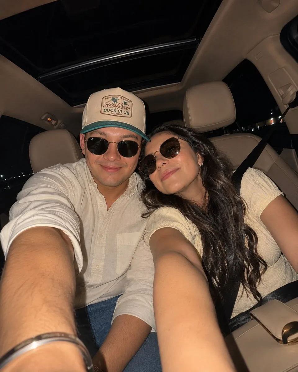 Warm car selfie shows a smiling couple wearing sunglasses in the back seat under soft flash lighting