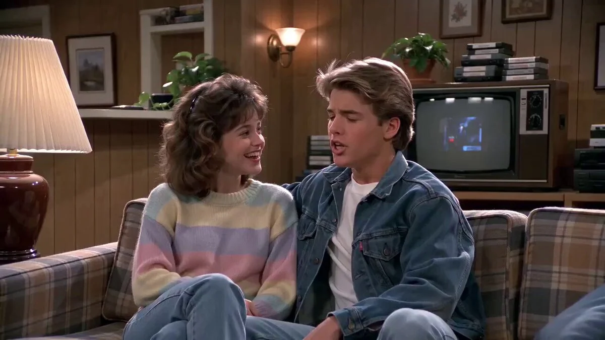 Two teenagers talk on a couch in a warm 1980s sitcom living room